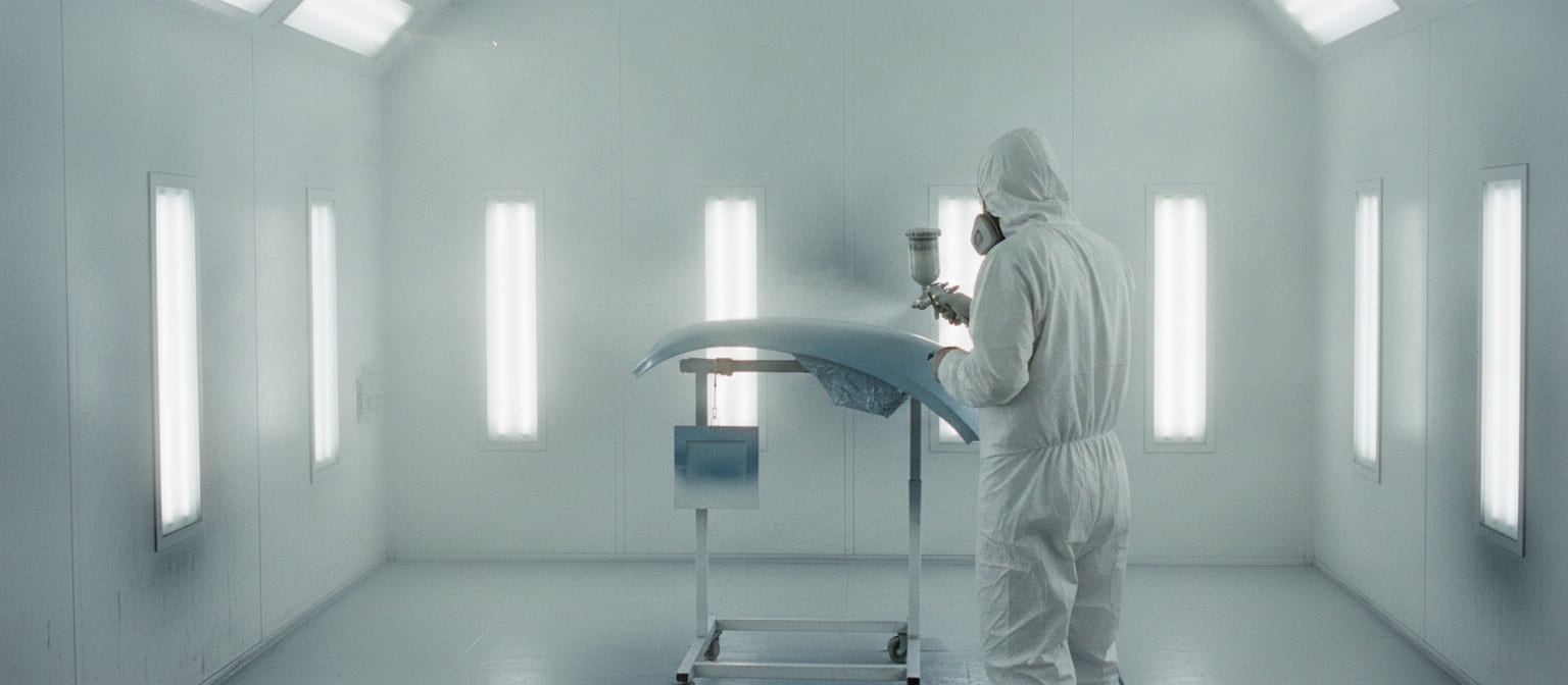 Inside a spotless downdraft paint booth: a painter in a white hooded suit and respirator sprays a pale blue-grey fender clamped to a rolling stand, under even white light from fixtures on every wall.