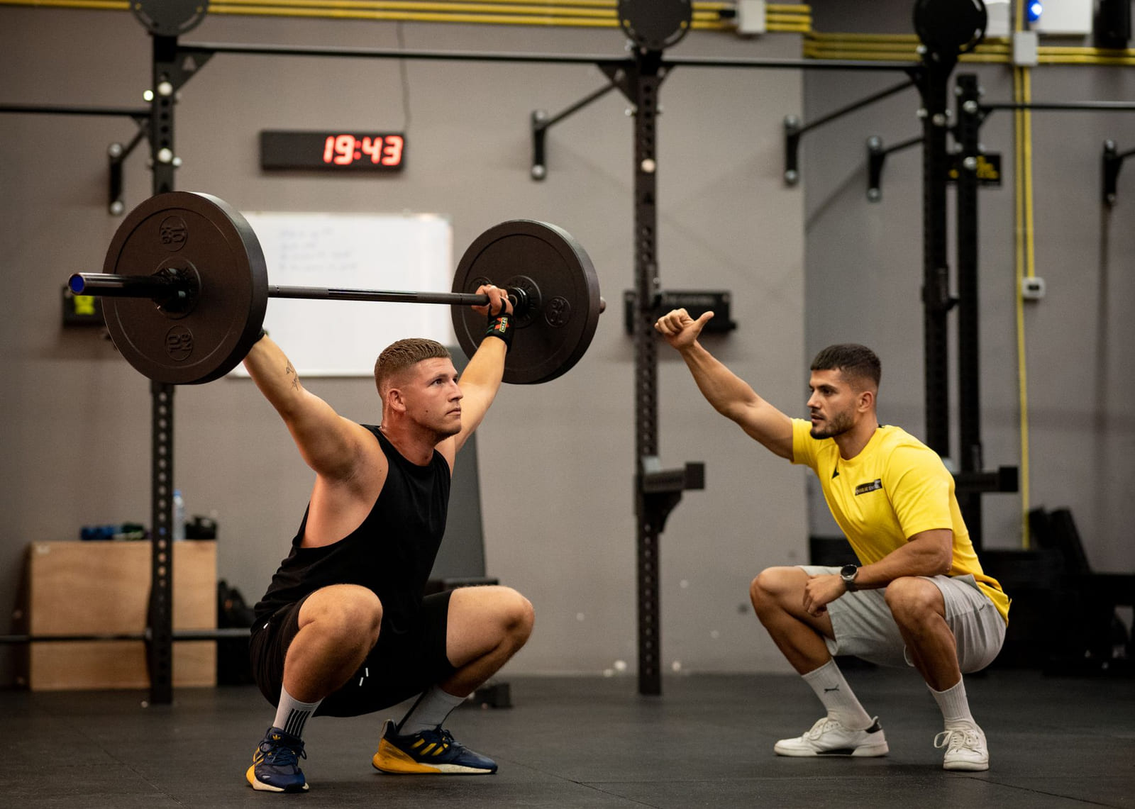 A coach standing beside a lifter working through a set with a loaded barbell.