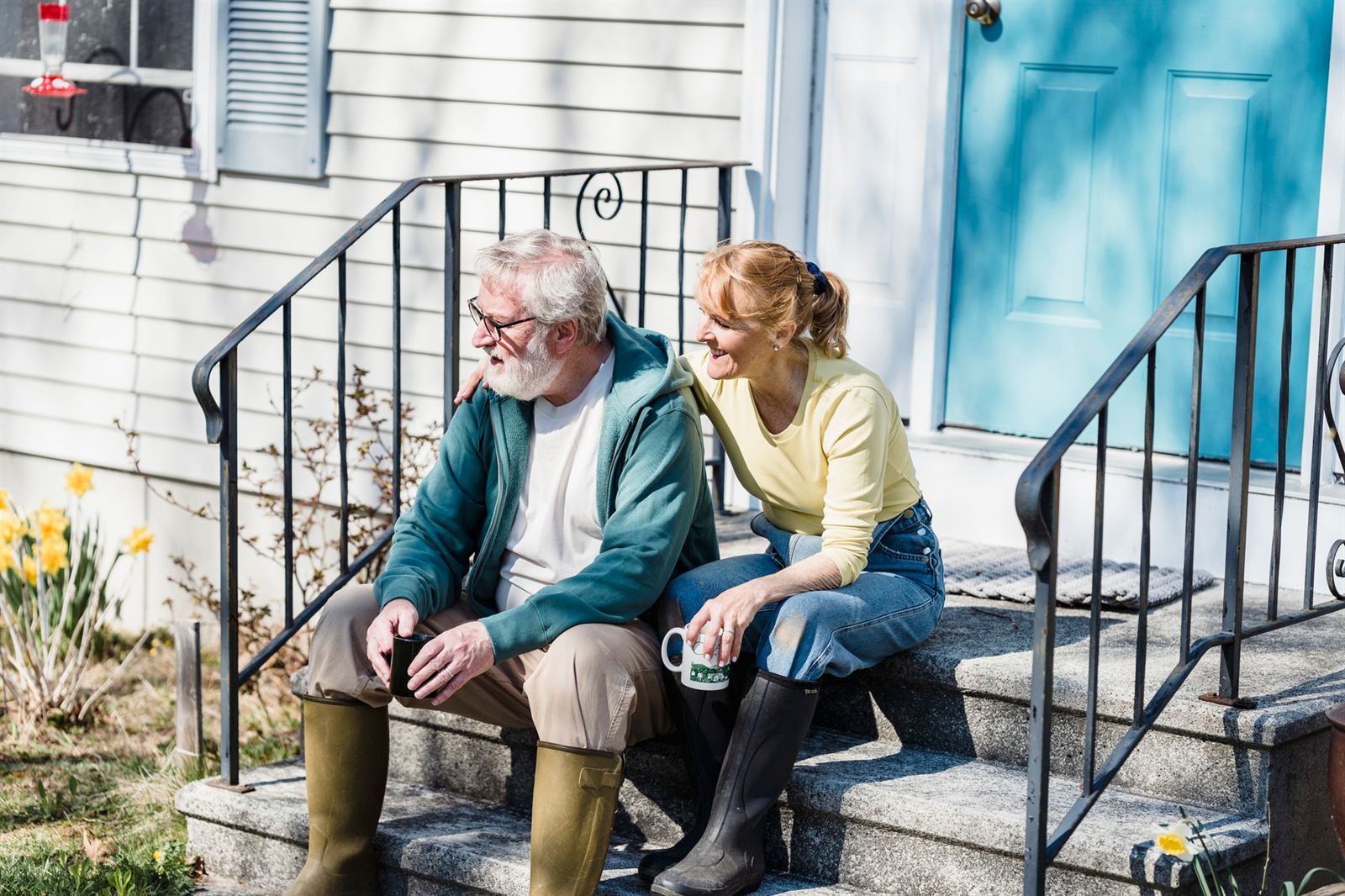 Two people sitting close on the front steps of a clapboard house with mugs of coffee, garden boots still on.
