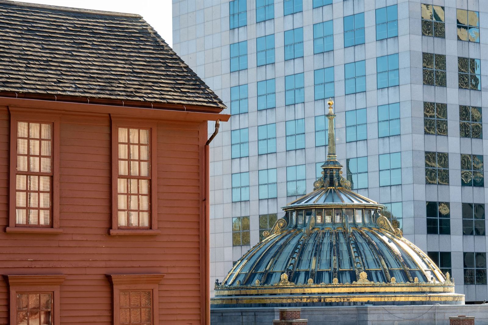 A red colonial house in Providence standing in front of a modern glass tower, with the gilded dome of an old bank between them.