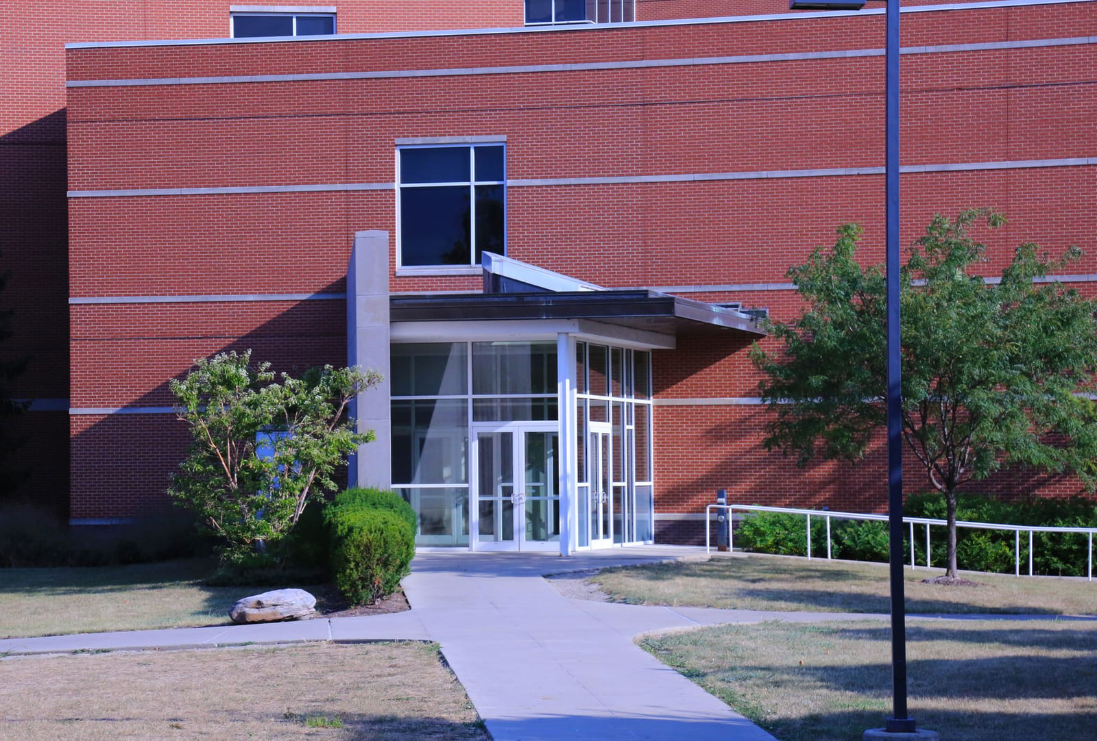 The glazed entrance of a low brick building with a shallow canopy, a concrete path and young trees on either side.