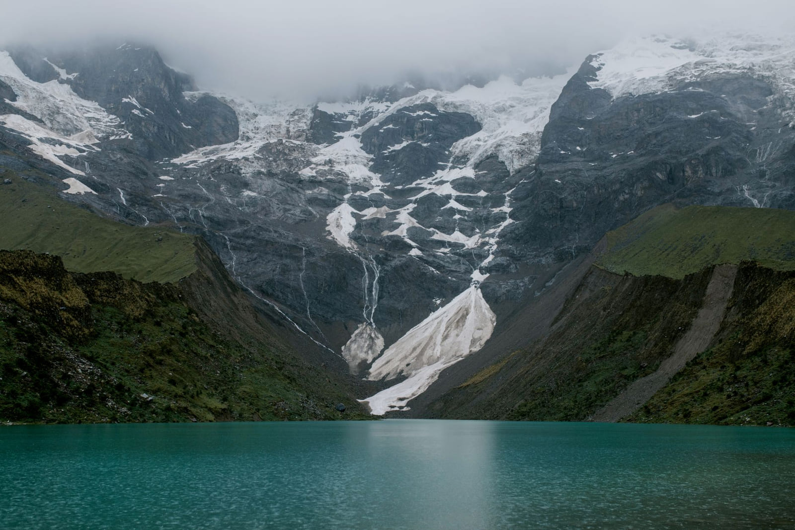 A turquoise glacier lake at the foot of a snow wall wrapped in mist