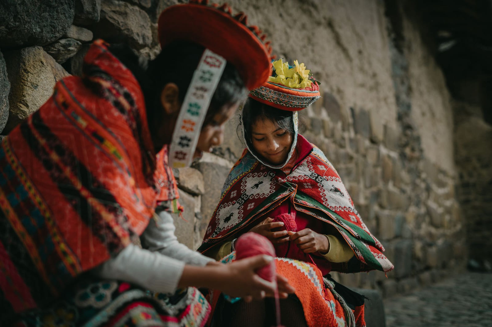 Two young weavers in traditional dress knit red yarn beside an Inca stone wall