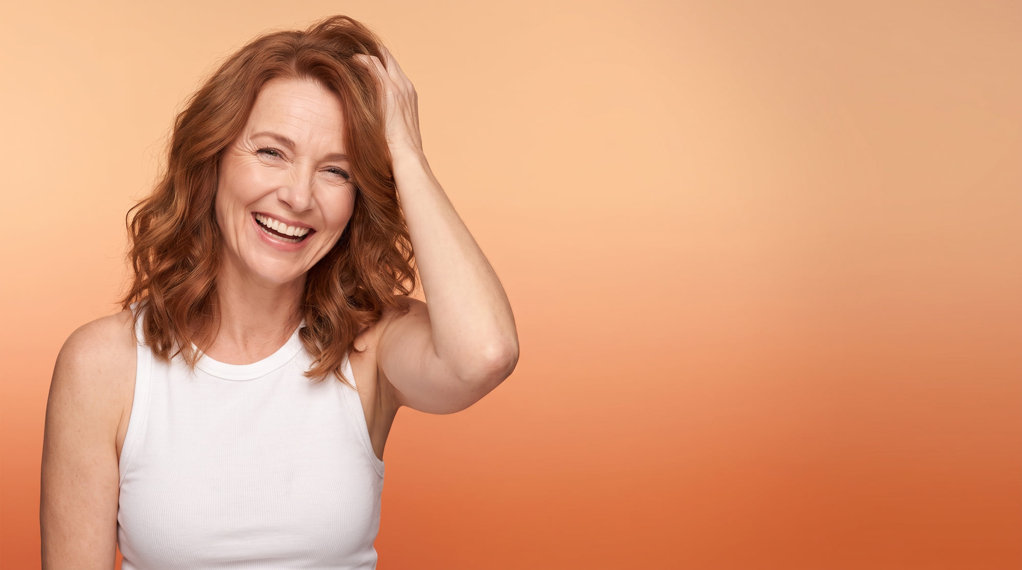 A woman in her fifties laughing with her eyebrows raised, on a peach background.