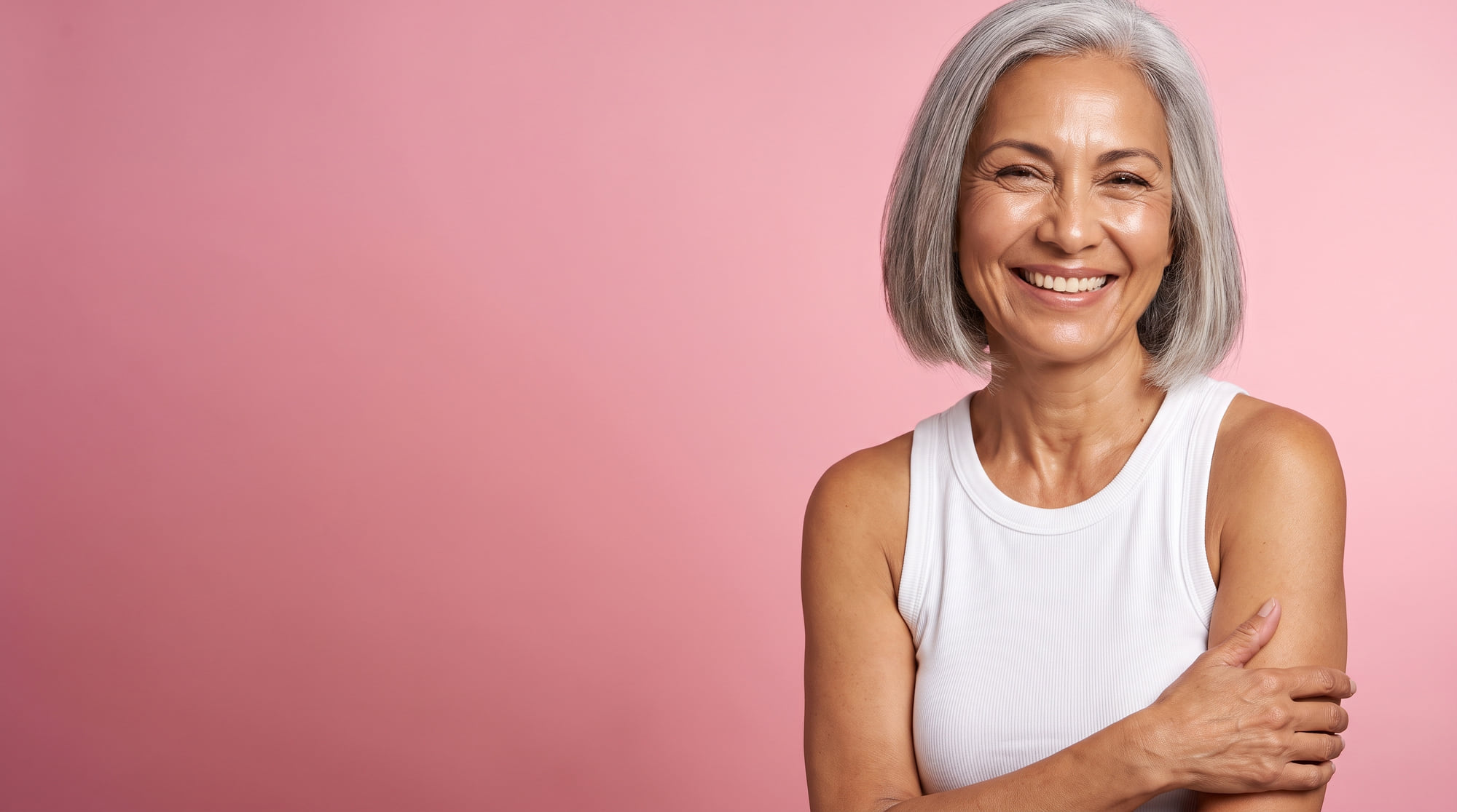 A woman in her sixties with silver hair smiling broadly, on a rose pink background.