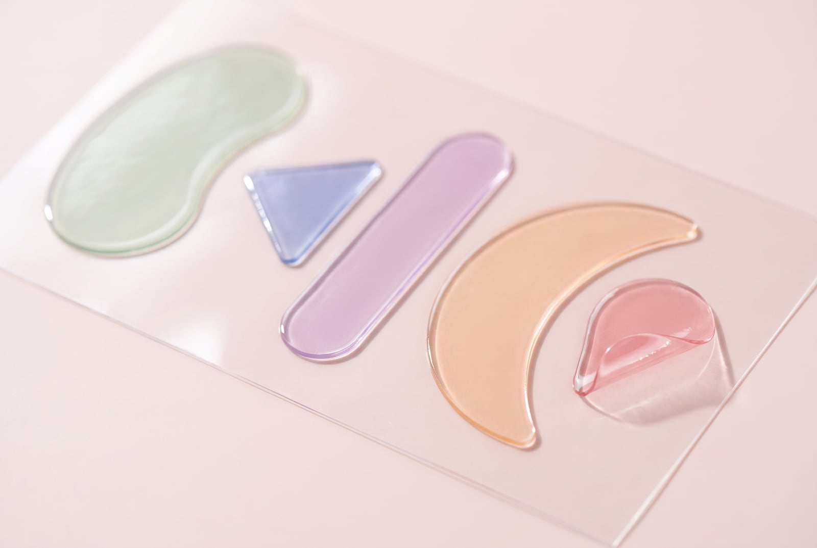 Five translucent facial patches on a clear backing sheet, one peeled up at the corner, each tinted a different pastel.