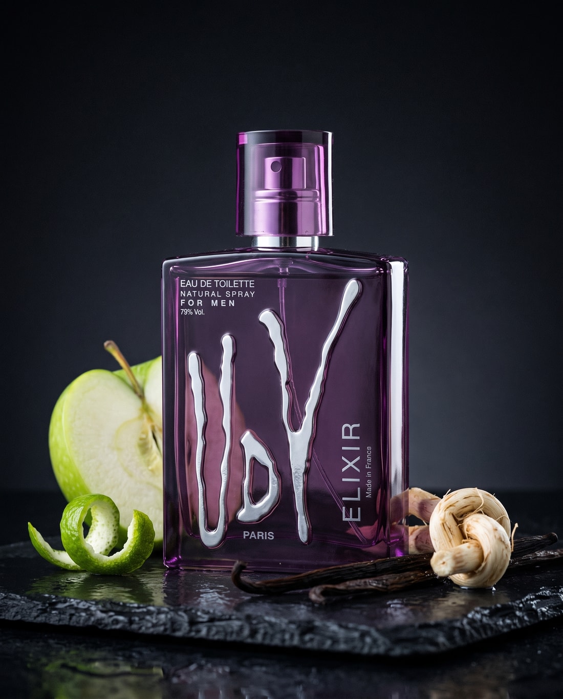 UDV Elixir beside a halved apple, bergamot peel, vanilla pods and vetiver root on wet slate.