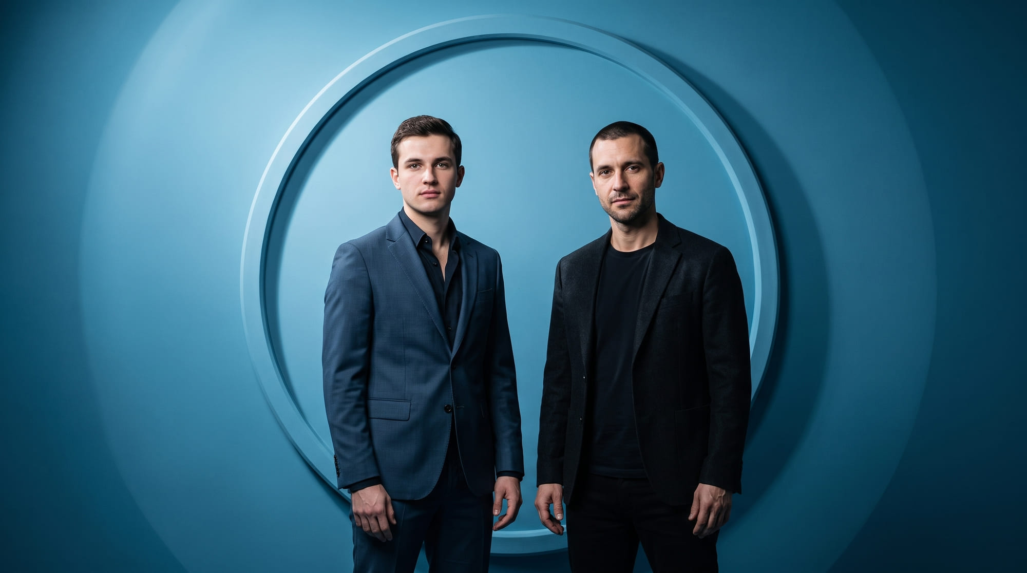 Two men in tailored jackets against a steel-cyan backdrop.
